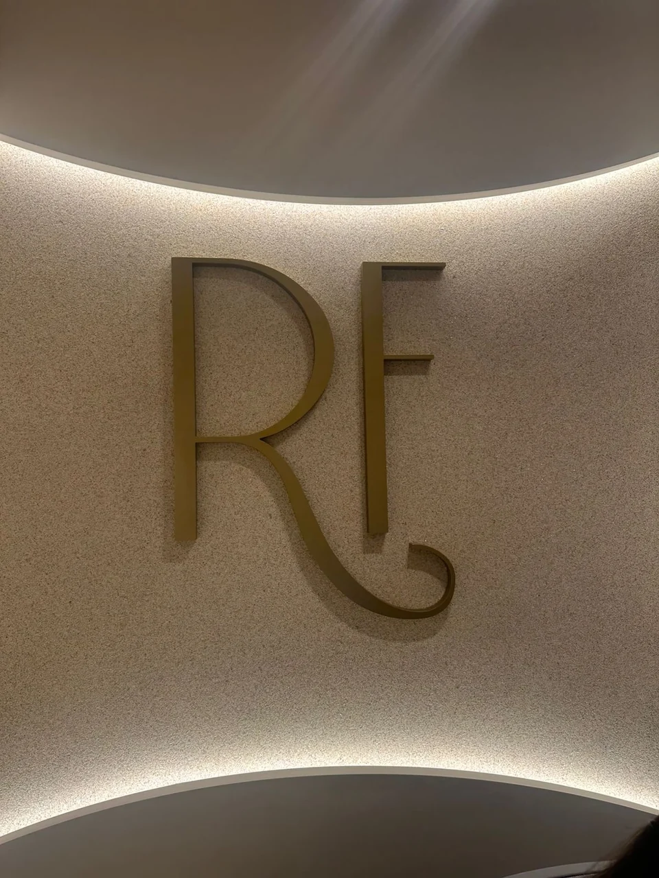 Monograma RF dourado em parede de textura clara com iluminação curva